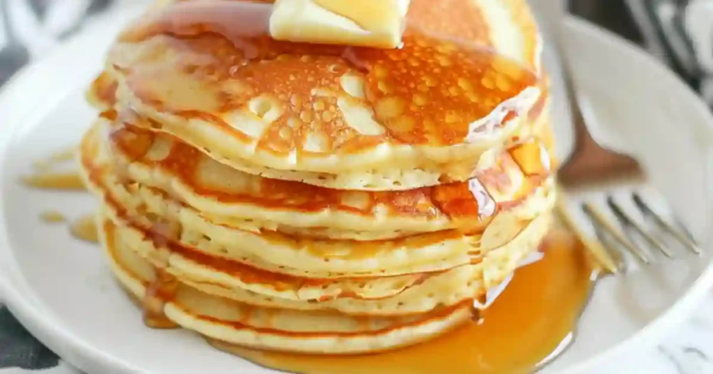 A stack of fluffy pancakes topped with a pat of butter and drizzled with syrup, served on a white plate.