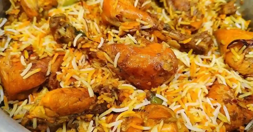 A close-up view of a pot filled with aromatic biryani, featuring golden pieces of chicken interspersed with vibrant, fluffy basmati rice garnished with orange and green spices.