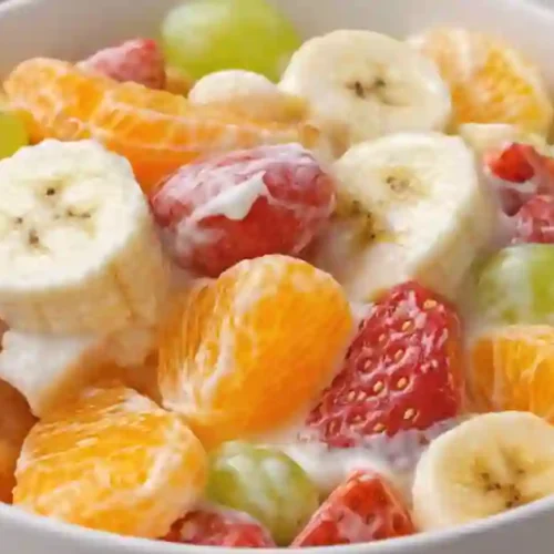 A bowl of creamy fresh fruit salad featuring banana, strawberries, green grapes, and tangerines, drizzled with yogurt dressing.