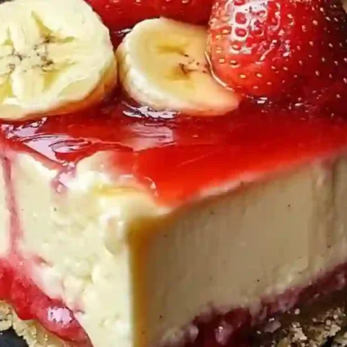 A close-up, macro shot of a thick square slice of cheesecake served on a dark plate. The dessert features a golden graham cracker crust at the bottom, a vibrant red strawberry jam layer, a thick and creamy pale-yellow cheesecake filling, and a glossy red strawberry glaze on top. It is garnished with fresh, round banana slices and a whole ripe strawberry.