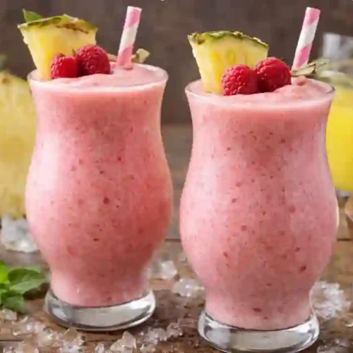 Two frozen Raspberry Piña Colada drinks garnished with pineapple and raspberries, surrounded by fresh raspberries and coconut cream.
