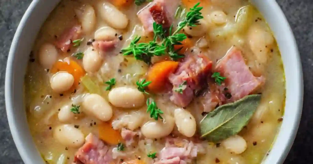 A warm bowl of white bean and meat soup, filled with chunks of meat, white beans, and colorful vegetables garnished with herbs.