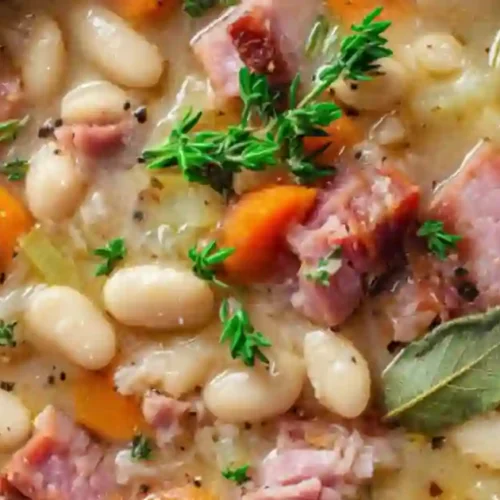 A warm bowl of white bean and meat soup, filled with chunks of meat, white beans, and colorful vegetables garnished with herbs.
