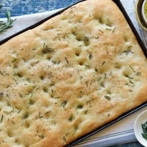 A beautifully risen overnight focaccia bread, golden brown and topped with fresh herbs, resting on a wooden board with a drizzle of olive oil nearby.