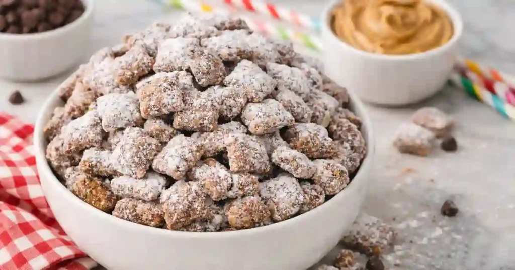 A bowl overflowing with homemade Muddy Buddies, dusted with powdered sugar, surrounded by bowls of chocolate chips and peanut butter, and festive decorations.