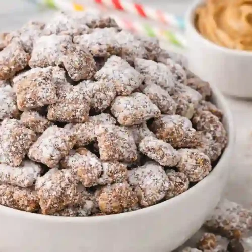 A bowl overflowing with homemade Muddy Buddies, dusted with powdered sugar, surrounded by bowls of chocolate chips and peanut butter, and festive decorations.