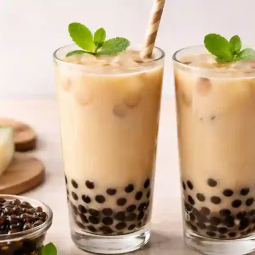 Two tall glasses of iced wintermelon milk tea with black tapioca pearls settled at the bottom, garnished with mint leaves and striped paper straws; sliced wintermelon, brown sugar, brewed tea, and a bowl of cooked boba sit on the counter in the background.