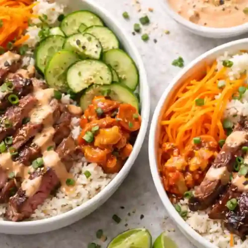 Two bowls of Korean BBQ steak rice topped with spicy cream sauce, cucumber slices, shredded carrots, and garnished with green onions, served with lime wedges.