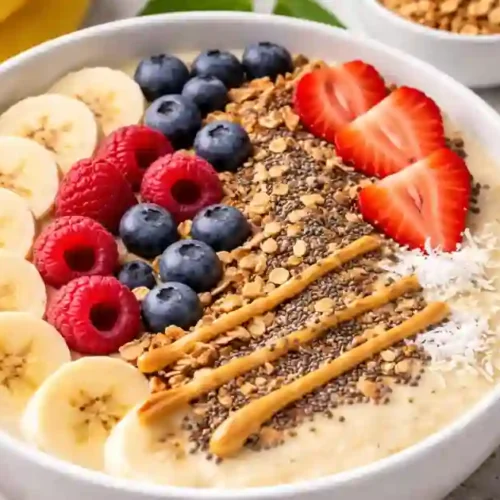 A top-down view of a vibrant white bowl filled with a thick banana smoothie base. The bowl is beautifully decorated with neat rows of sliced bananas, fresh blueberries, raspberries, and sliced strawberries. It is also topped with crunchy granola, chia seeds, shredded coconut, and a drizzle of creamy peanut butter. Fresh whole bananas and strawberries are scattered on the light grey stone background.