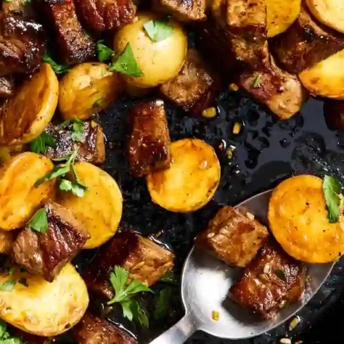 Top-down view of a cast-iron skillet filled with browned garlic butter steak bites and halved golden baby potatoes. The steak pieces are seared with a caramelized crust, the potatoes are crispy and seasoned, and small parsley leaves are scattered on top. A spoon lifts a piece of steak and a potato showing the juicy interior.