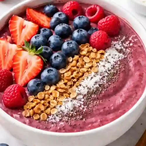 A top-down, slightly angled view of a thick purple berry smoothie bowl in a white ceramic dish. The bowl is topped with neat rows of sliced strawberries, fresh blueberries, raspberries, crunchy granola, shredded coconut, and chia seeds. A wooden spoon rests beside the bowl on a white marble surface.