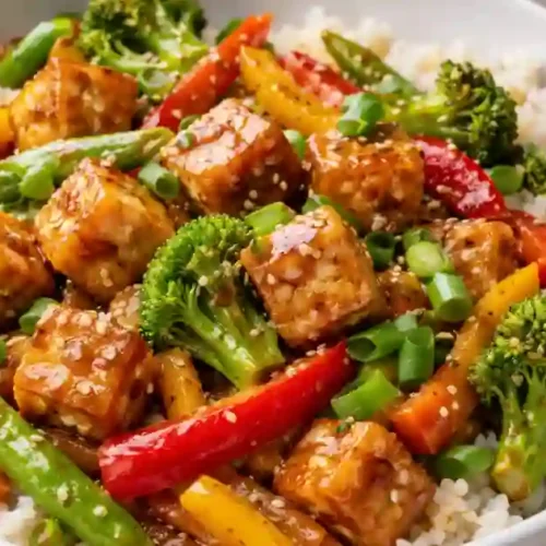 A white bowl filled with a colorful tofu stir fry served over a bed of white rice. The dish features golden-brown crispy tofu cubes, broccoli florets, sliced red and yellow bell peppers, and snap peas, all coated in a glossy sauce and garnished with sesame seeds and chopped green onions. Wooden chopsticks rest on the edge of the bowl.