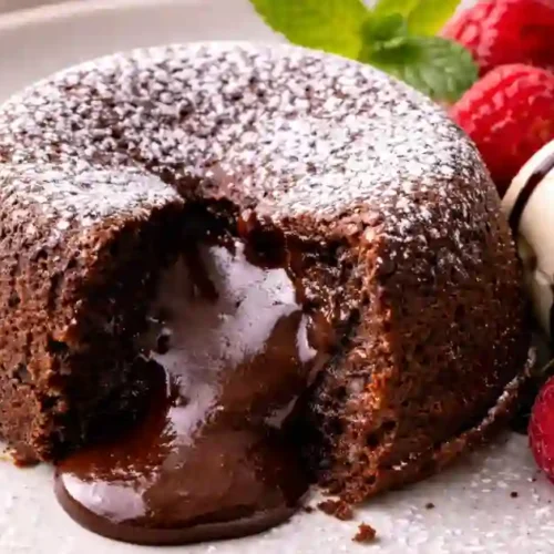 A close-up of a warm molten chocolate lava cake on a white plate, with its gooey chocolate center flowing out. The cake is dusted with powdered sugar and served with a scoop of vanilla ice cream drizzled with chocolate syrup, fresh raspberries, and a mint leaf garnish.