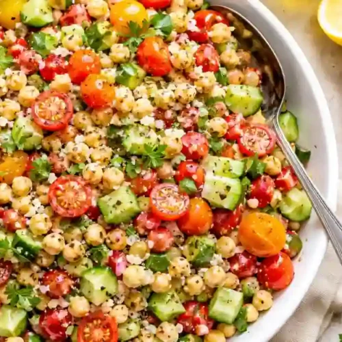 A large white bowl filled with a colorful chickpea salad. The salad features cooked chickpeas, halved cherry tomatoes, diced cucumbers, and fresh parsley, all tossed in a light herb dressing. A silver serving spoon is in the bowl, and small side dishes of feta cheese, kalamata olives, whole garlic, and dried oregano are visible on a light-colored stone surface.