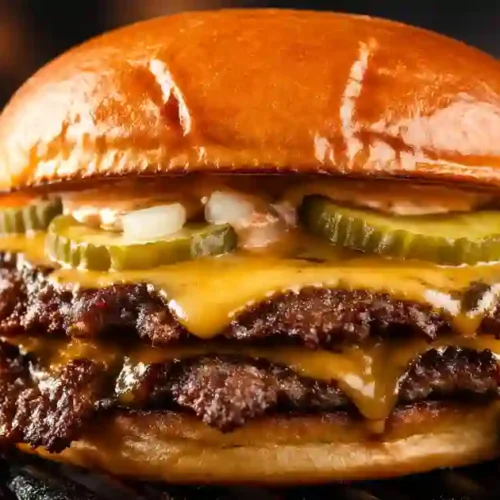 A close-up of a double smashed cheeseburger on a grill. The burger features two thin beef patties with jagged, crispy edges, topped with melted yellow cheese, crinkle-cut pickles, diced white onions, and a creamy sauce inside a toasted brioche bun. Flames from the grill are visible in the blurred background.