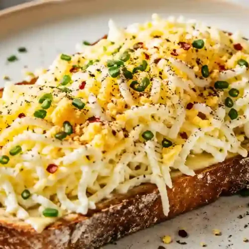 A close-up shot of a thick slice of toasted sourdough bread topped with a mountain of finely grated hard-boiled egg. The dish is garnished with fresh chopped chives, red pepper flakes, and black pepper.
