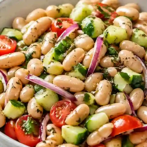 A close-up overhead view of a large white bowl filled with a colorful bean salad. The dish features plump white cannellini beans mixed with halved cherry tomatoes, diced cucumbers, and thinly sliced red onions. The salad is tossed in a herb-flecked dressing and garnished with fresh parsley.