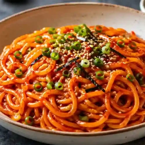 A close-up shot of a large bowl filled with thick, chewy noodles coated in a vibrant, glossy red Gochujang sauce. The noodles are garnished with sliced green onions, sesame seeds, and small strips of dried seaweed. A pair of wooden chopsticks is lifting a portion of the noodles, with small bowls of extra sauce and garnish visible in the background.
