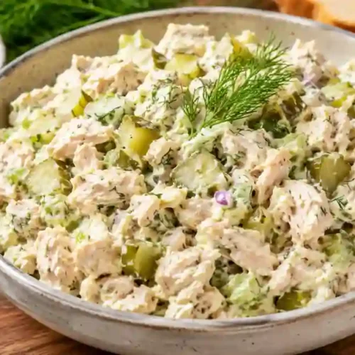 A close-up shot of a large ceramic bowl filled with creamy chicken salad. The salad features shredded chicken mixed with diced dill pickles, celery, and fresh dill. A sprig of fresh dill sits on top as a garnish. In the background, a small side bowl of pickle slices and pieces of crusty bread are visible on a wooden surface.