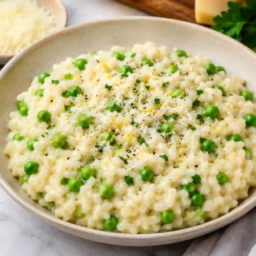 A close-up shot of a creamy white risotto studded with bright green peas, served in a shallow beige ceramic bowl on a marble surface. The dish is finished with freshly grated parmesan cheese, cracked black pepper, and fine lemon zest. In the background, a small bowl of grated parmesan, a wooden bowl of black peppercorns, fresh parsley sprigs, a block of cheese, and a lemon half are neatly arranged on a wooden board.