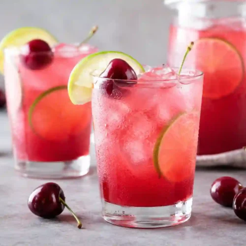 Two tall, condensation-covered glasses and a large glass pitcher in the background are filled with a vibrant pink-red cherry limeade over plenty of crushed ice. Each glass is elegantly garnished with a round lime wheel tucked inside, a fresh lime wedge on the rim, and a whole dark red sweet cherry with its stem. Scattered around the glasses on a grey concrete-textured surface are several whole fresh cherries with stems, a halved lime, and a small bowl of cherries blurred out in the background.