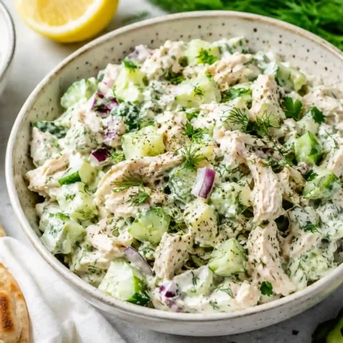 A large ceramic bowl filled with creamy Tzatziki chicken salad, featuring shredded chicken breast, diced English cucumbers, and finely chopped red onion tossed in a thick, herb-flecked Greek yogurt dressing. The salad is garnished with cracked black pepper, fresh dill sprigs, and flat-leaf parsley. In the background, warm flatbread or pita sits on a white cloth alongside a small bowl of extra tzatziki sauce, a cut lemon half, a bunch of fresh dill, and sliced cucumbers on a light gray countertop.
