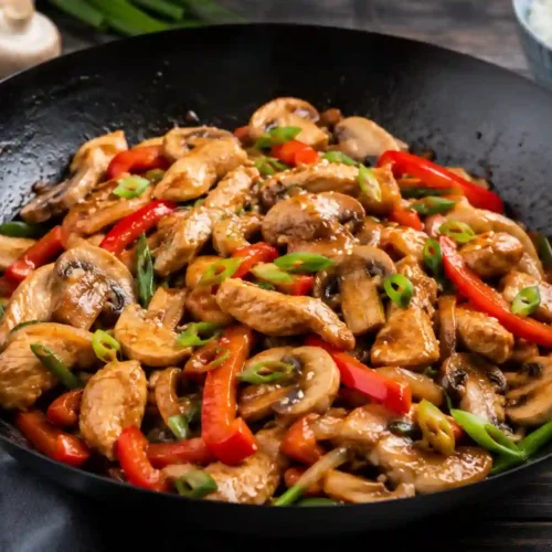 A close-up, high-angle shot of a classic black carbon steel wok filled with a glossy chicken and mushroom stir fry. The dish features tender, golden-brown strips of chicken breast, thick-sliced white button mushrooms, and vibrant red bell pepper strips, all coated in a rich, shimmering brown savory sauce. The stir fry is heavily garnished with freshly chopped green onions. In the background, a dark wooden surface hosts a blurred blue ceramic bowl filled with fluffy white rice, fresh whole mushrooms, and a bundle of green onions on top of a dark gray cloth napkin.