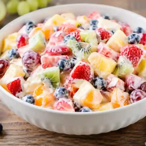A large, white ribbed ceramic bowl overflowing with a colorful, creamy fruit salad on a rustic wooden tabletop. The salad features chopped strawberries, cubed pineapple, sliced kiwi, fresh blueberries, mandarin orange segments, and whole red grapes, all tossed in a thin, glossy, semi-translucent creamy dressing. Fresh strawberries, blueberries, green grapes, and a halved kiwi are scattered around the base of the bowl on the wooden surface, alongside a folded white kitchen towel with gray stripes.