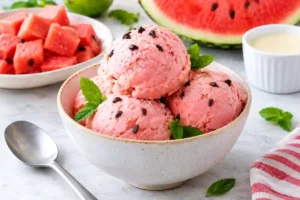 A ceramic bowl filled with four round scoops of light pink watermelon ice cream dotted with mini dark chocolate chips and garnished with fresh mint leaves. The bowl sits on a white marble surface next to a spoon, a red-striped napkin, a bowl of diced watermelon cubes, a slice of fresh watermelon, a small dish of sweetened condensed milk, and fresh mint sprigs in the soft-focused background.