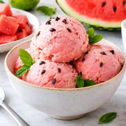 A ceramic bowl filled with four round scoops of light pink watermelon ice cream dotted with mini dark chocolate chips and garnished with fresh mint leaves. The bowl sits on a white marble surface next to a spoon, a red-striped napkin, a bowl of diced watermelon cubes, a slice of fresh watermelon, a small dish of sweetened condensed milk, and fresh mint sprigs in the soft-focused background.